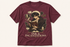 A maroon short-sleeve graphic t-shirt featuring an illustration of a young man reading a book surrounded by ink, quills, and cards, with the text &