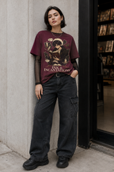 A model stands against a concrete wall wearing a maroon graphic t-shirt with a gothic illustration and the text &