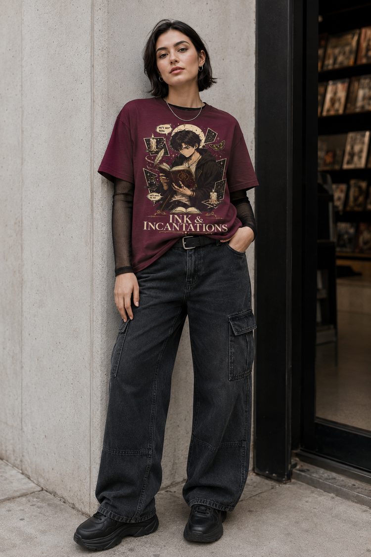 A model stands against a concrete wall wearing a maroon graphic t-shirt with a gothic illustration and the text &