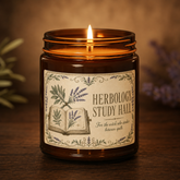 A lit amber glass jar candle on a dark wooden surface with a warm blurred background. The candle has a cream label with botanical illustrations and the text &