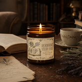 A lit amber glass jar candle on a rustic wooden table, surrounded by an open book, a cup and saucer, and dried lavender. The candle label reads &