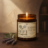 An amber glass jar candle with a lit wick sits on a wooden surface, styled with dried lavender sprigs. The label reads &