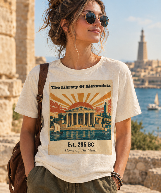 A woman wearing round sunglasses and a cream-colored graphic t-shirt with a vintage-style print of The Library of Alexandria, standing outdoors near ancient stone ruins and the sea.