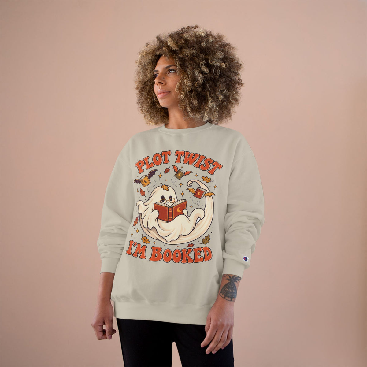 A woman with curly hair models a light beige crewneck sweatshirt featuring a cartoon ghost reading a book with the text &