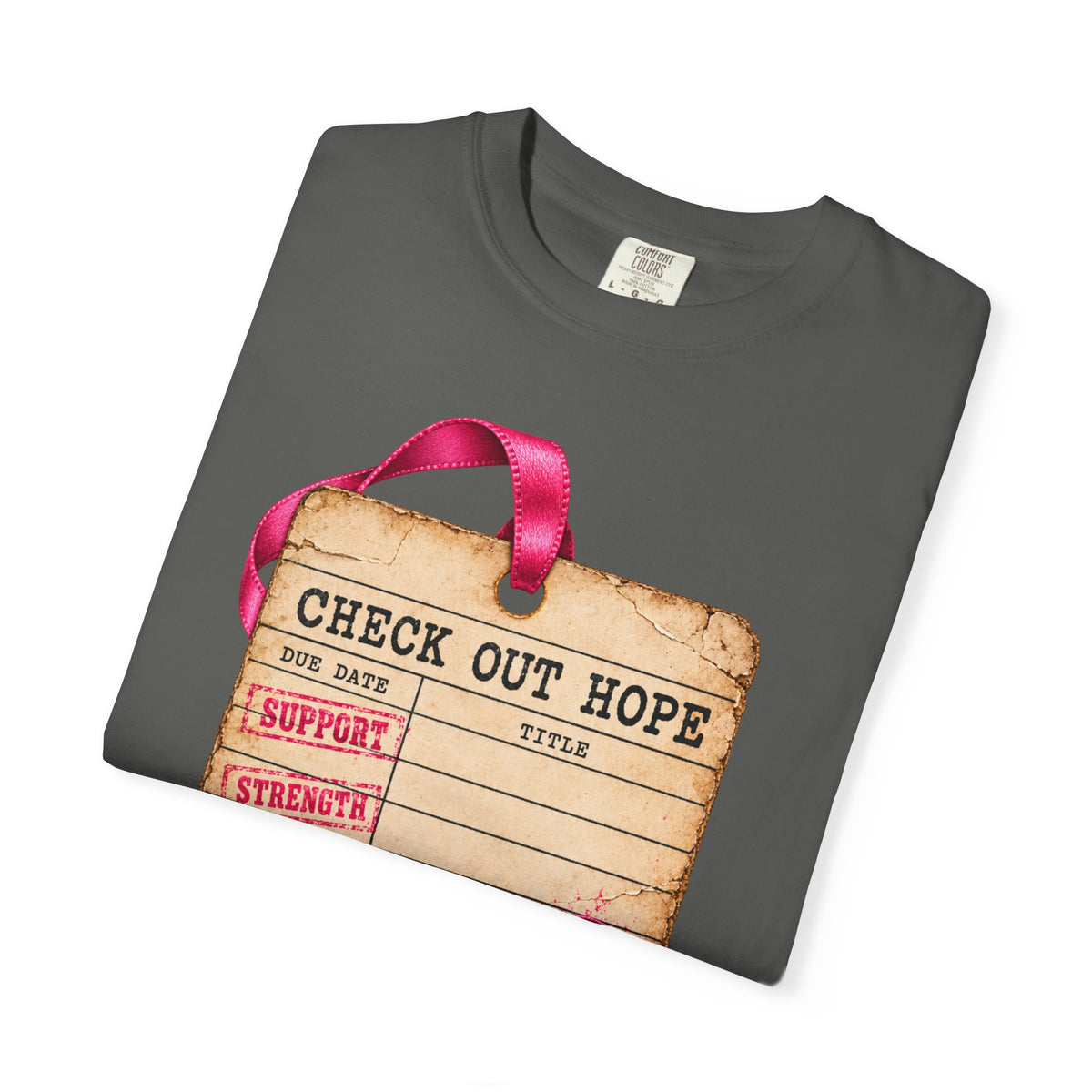 A dark gray folded t-shirt featuring a graphic print of a vintage luggage tag with a pink ribbon handle. The tag has a distressed beige background with black text reading &