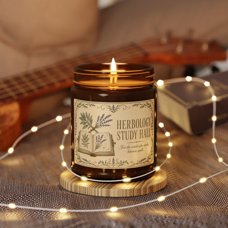 An amber glass jar candle with a lit wick sits on a small wooden coaster, surrounded by warm fairy lights. The label reads &
