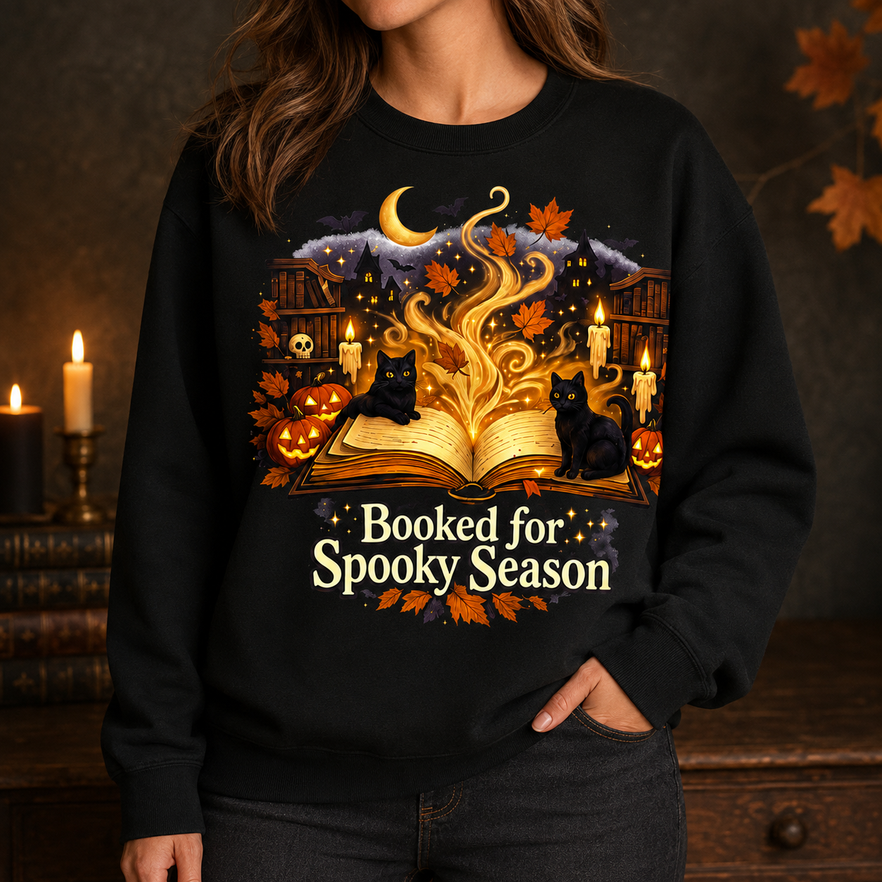 A woman wearing a black crewneck sweatshirt with a detailed Halloween graphic. The graphic features an open glowing book, two black cats, pumpkins, candles, autumn leaves, a crescent moon, and the text &