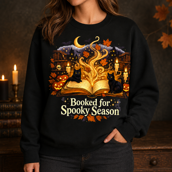 A woman wearing a black crewneck sweatshirt with a detailed Halloween graphic. The graphic features an open glowing book, two black cats, pumpkins, candles, autumn leaves, a crescent moon, and the text &