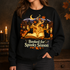 A woman wearing a black crewneck sweatshirt with a detailed Halloween graphic. The graphic features an open glowing book, two black cats, pumpkins, candles, autumn leaves, a crescent moon, and the text &