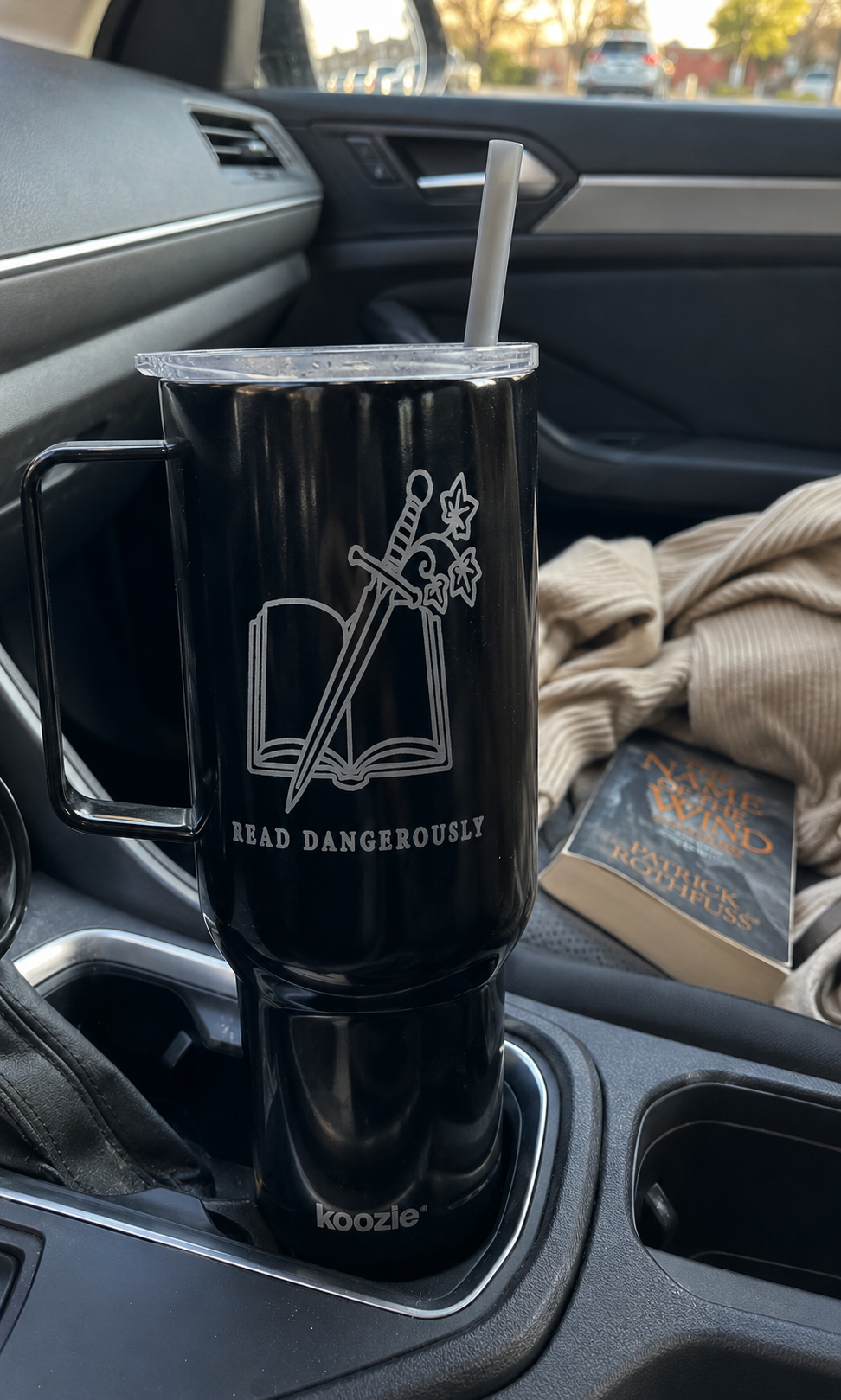 A black insulated travel tumbler with a handle and clear lid sits in a car cup holder. The tumbler has a white graphic of an open book with crossed swords and the text &