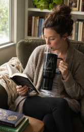 A woman in a cozy living room setting, wearing a beige cardigan and black pants, drinking from a black insulated tumbler with a straw while reading a book.