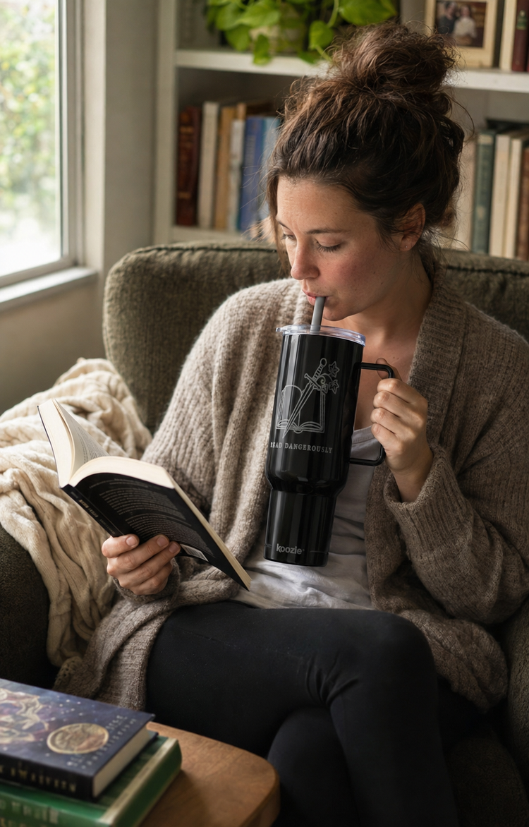 A woman in a cozy living room setting, wearing a beige cardigan and black pants, drinking from a black insulated tumbler with a straw while reading a book.