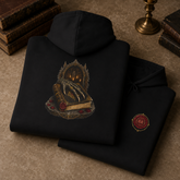 A black hoodie is folded and displayed on a textured brown surface with vintage books and a brass candlestick in the background. The hoodie features a detailed graphic of a skeleton key in an arched frame holding an open book with red wax seals, and a small red and gold seal graphic on the sleeve.