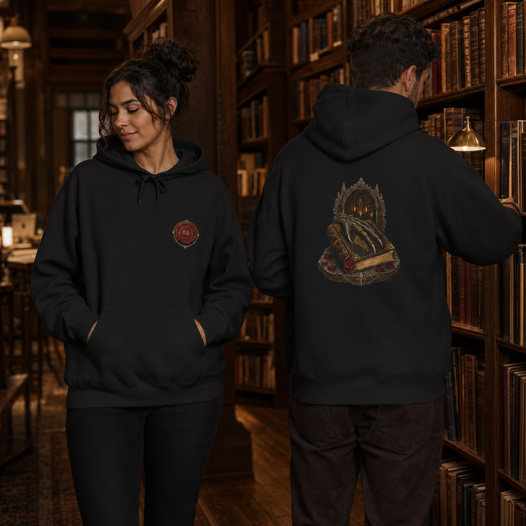 Two models wearing a black hoodie in a cozy, dimly lit library setting. The model on the left shows the front of the hoodie, which features a small circular red and gold crest on the left chest.